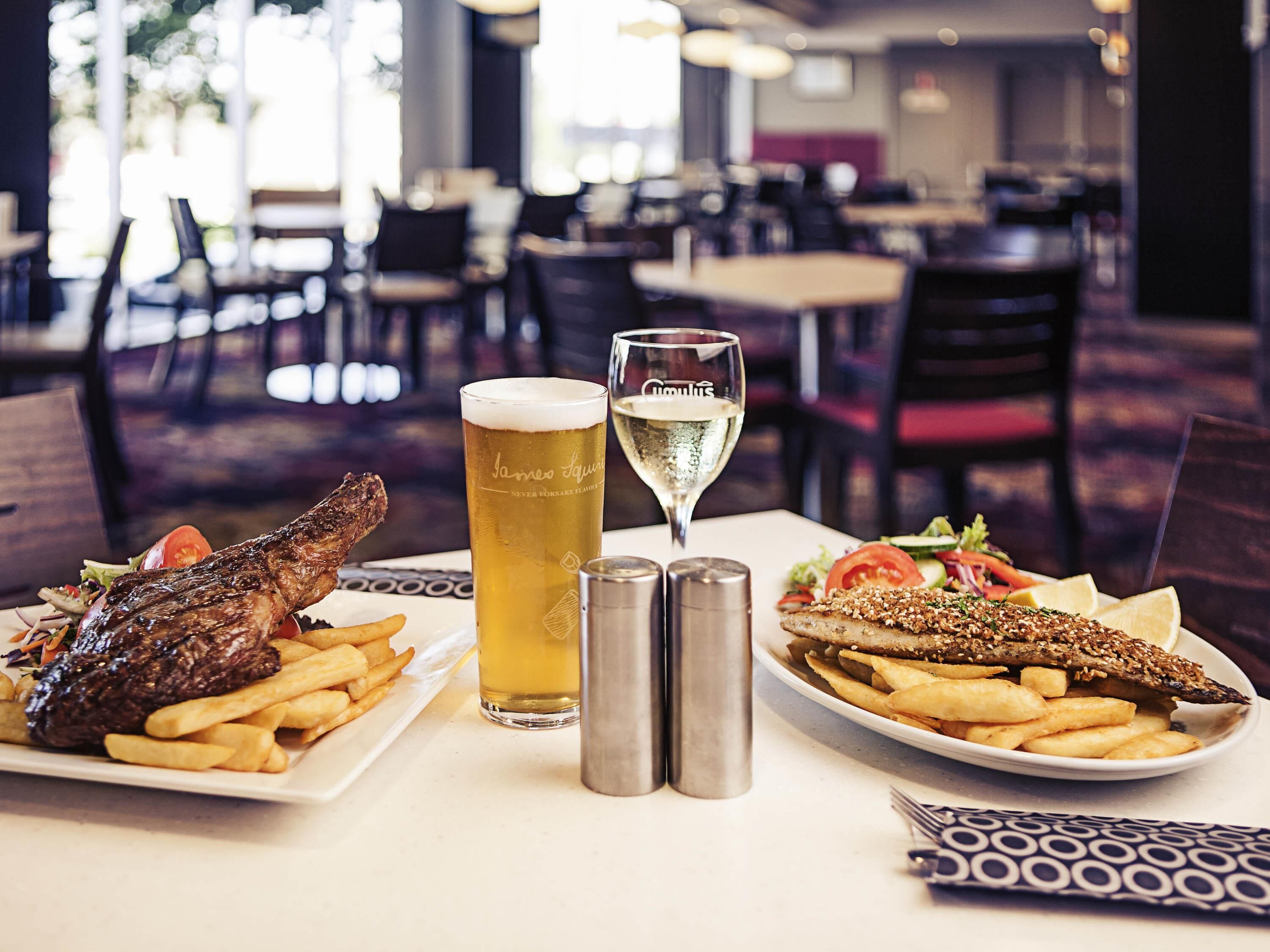 A restaurant setting with a table set for two, featuring a steak, fries, a fish dish, and a glass of beer.