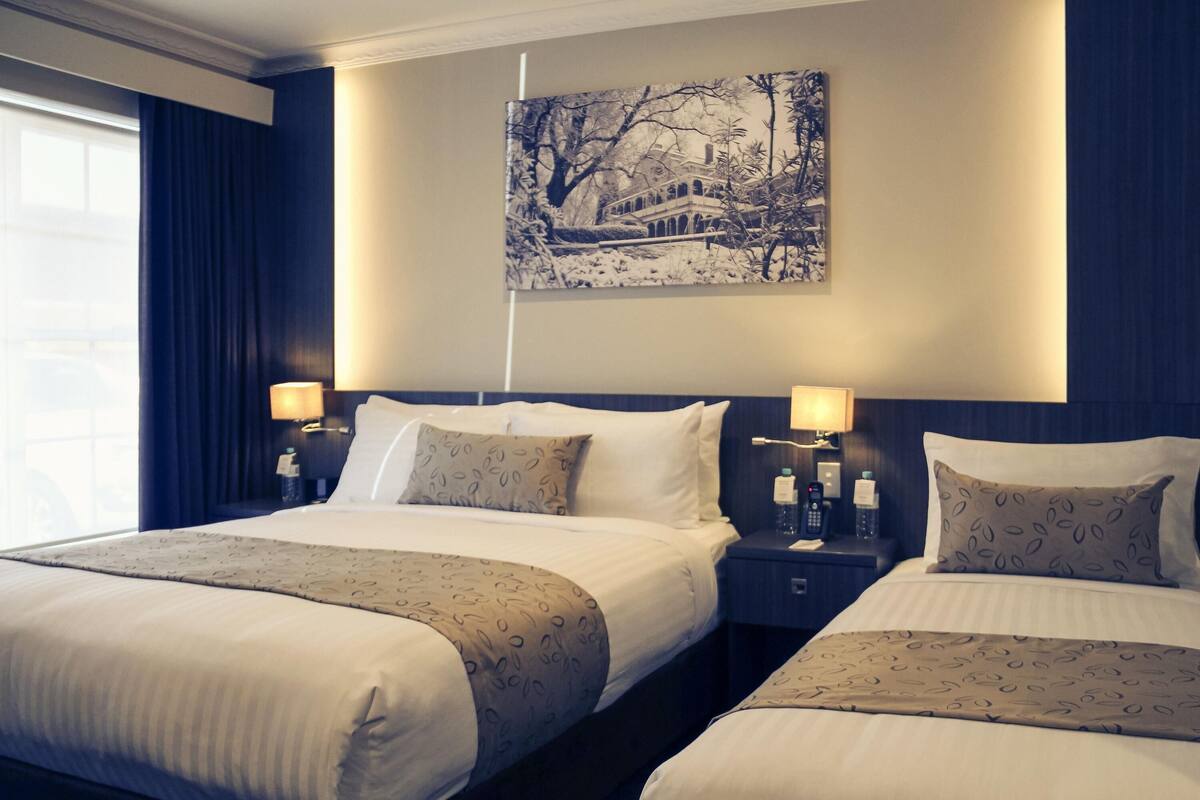 A hotel room with two beds, a nightstand with a lamp, a painting on the wall, and a window with curtains.
