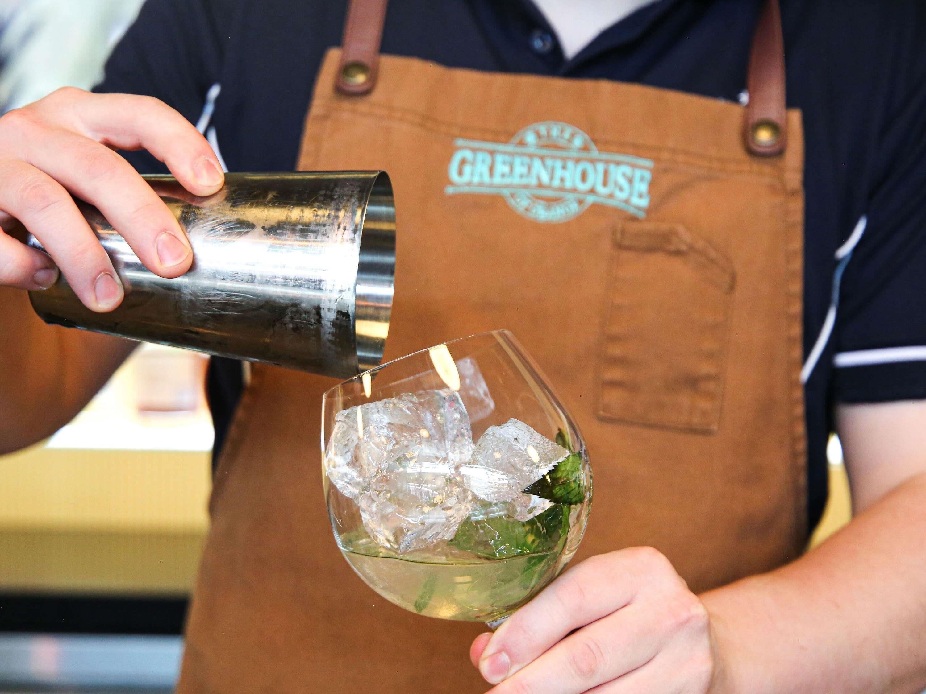 A person in a brown apron pouring a drink into a glass with ice.