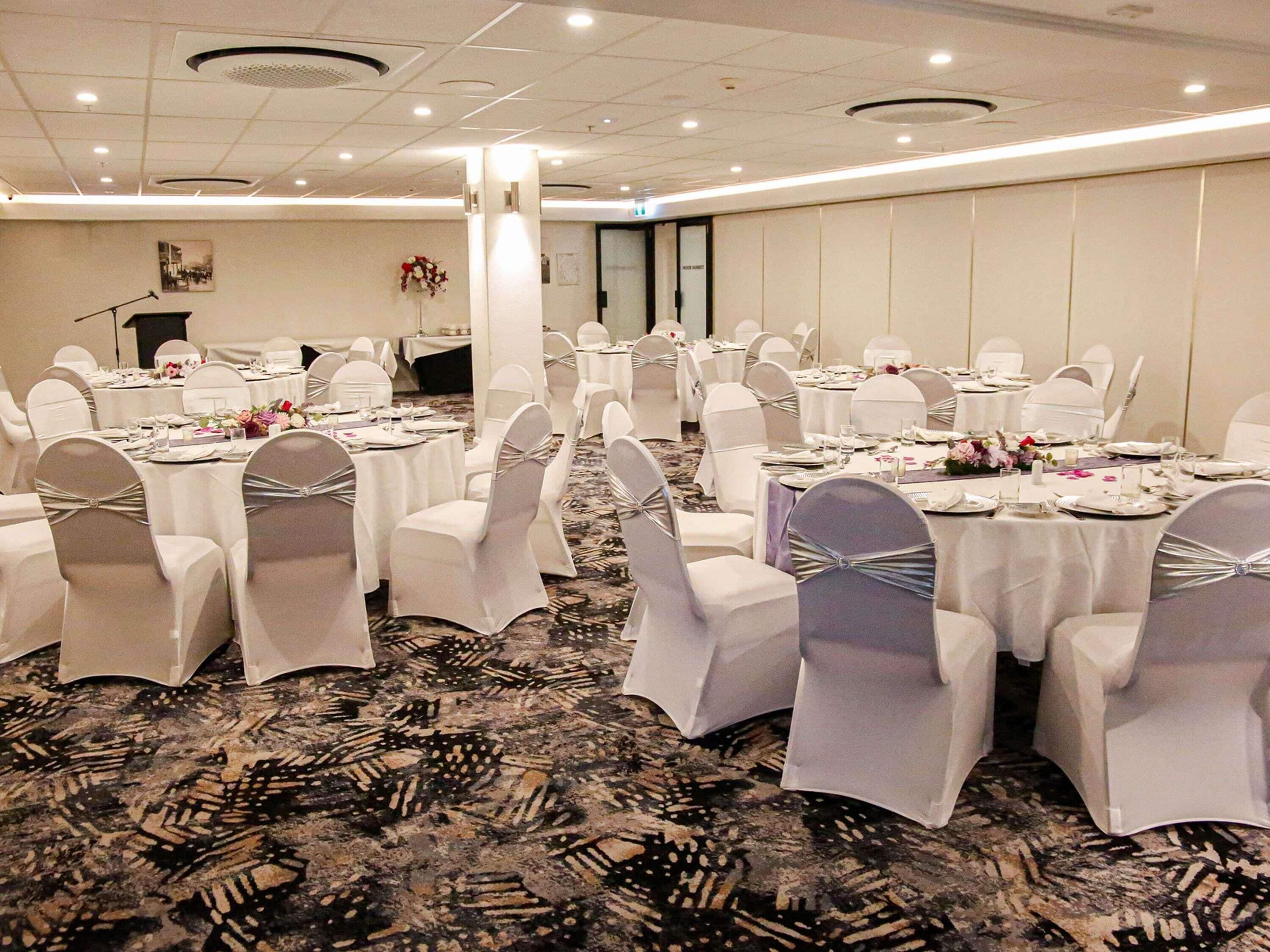 Un salón de banquetes con mesas redondas, sillas blancas y piso alfombrado.