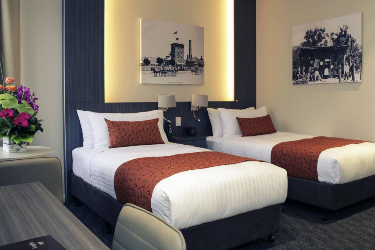 A hotel room with two beds, a nightstand, a lamp, a vase with flowers, and two wall-mounted pictures.