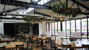 A spacious indoor venue with multiple round tables and chairs, large windows, and hanging floral arrangements. - Mercure Orange (Orange)