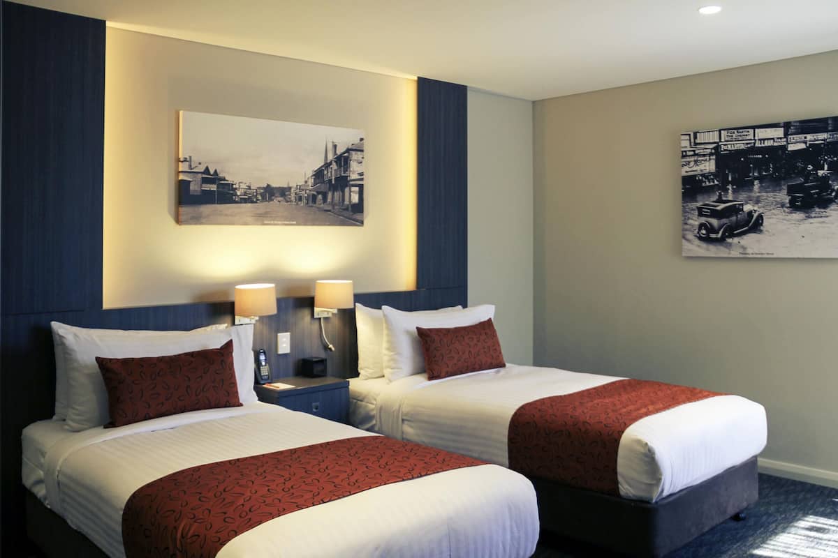 A hotel room with two beds, a nightstand with lamps, a wall-mounted painting, and a carpeted floor.