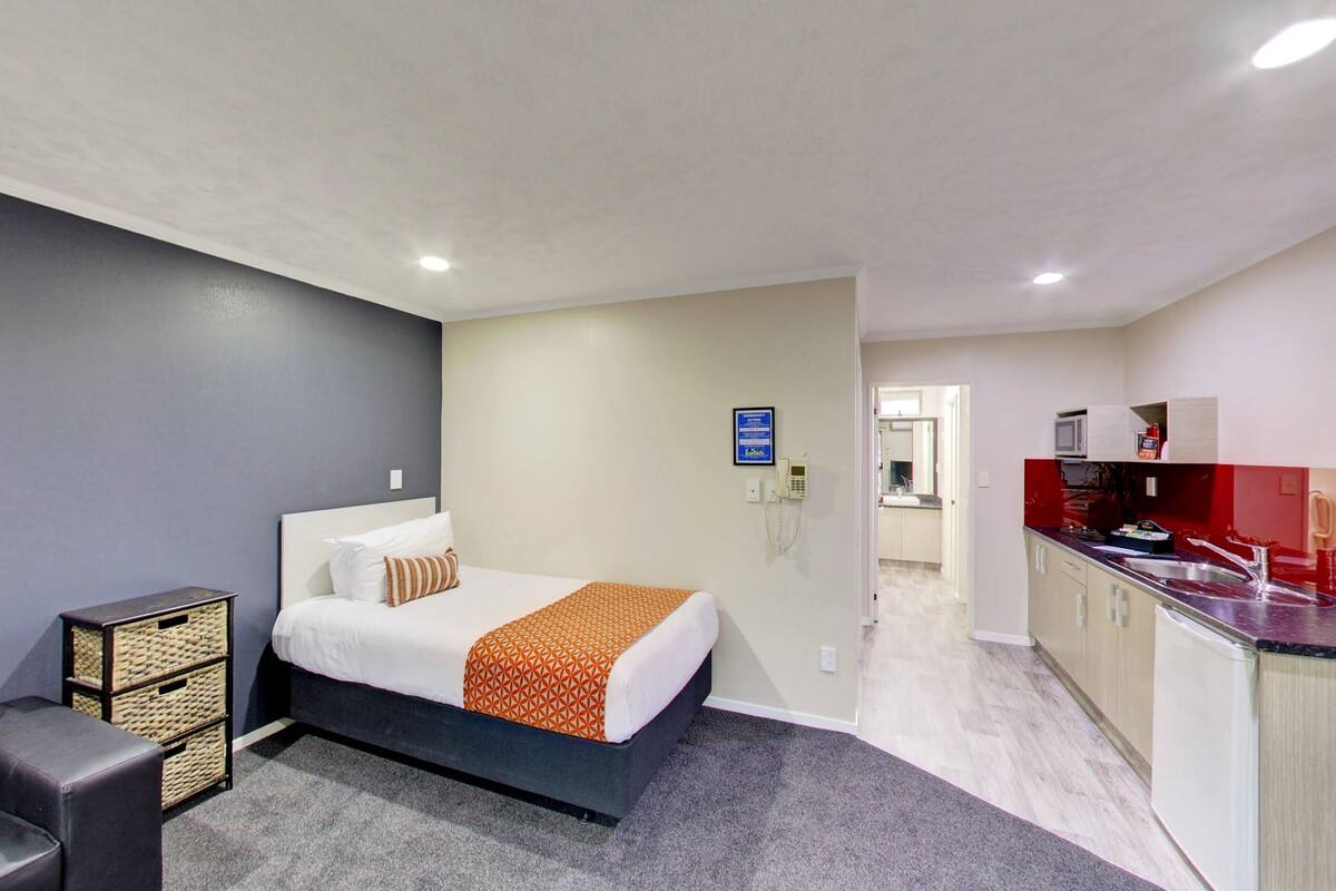 A hotel room with a bed, a small sofa, a kitchenette, and a bathroom visible through an open door.