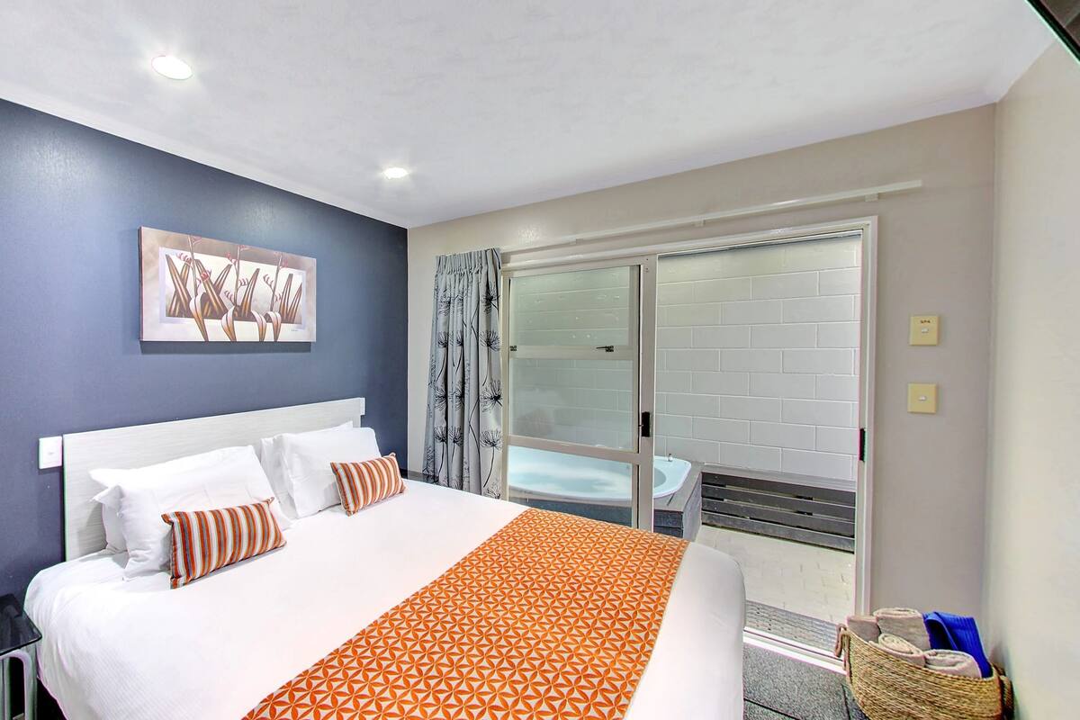 A hotel room with a large bed, a painting on the wall, a sliding glass door to a balcony, and a small table with a basket.