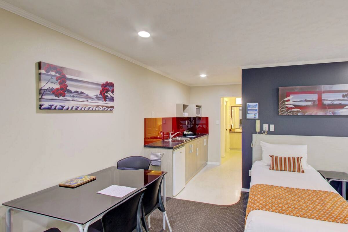 A hotel room with a bed, a desk, and a kitchenette. There is a painting on the wall and a window with a view.