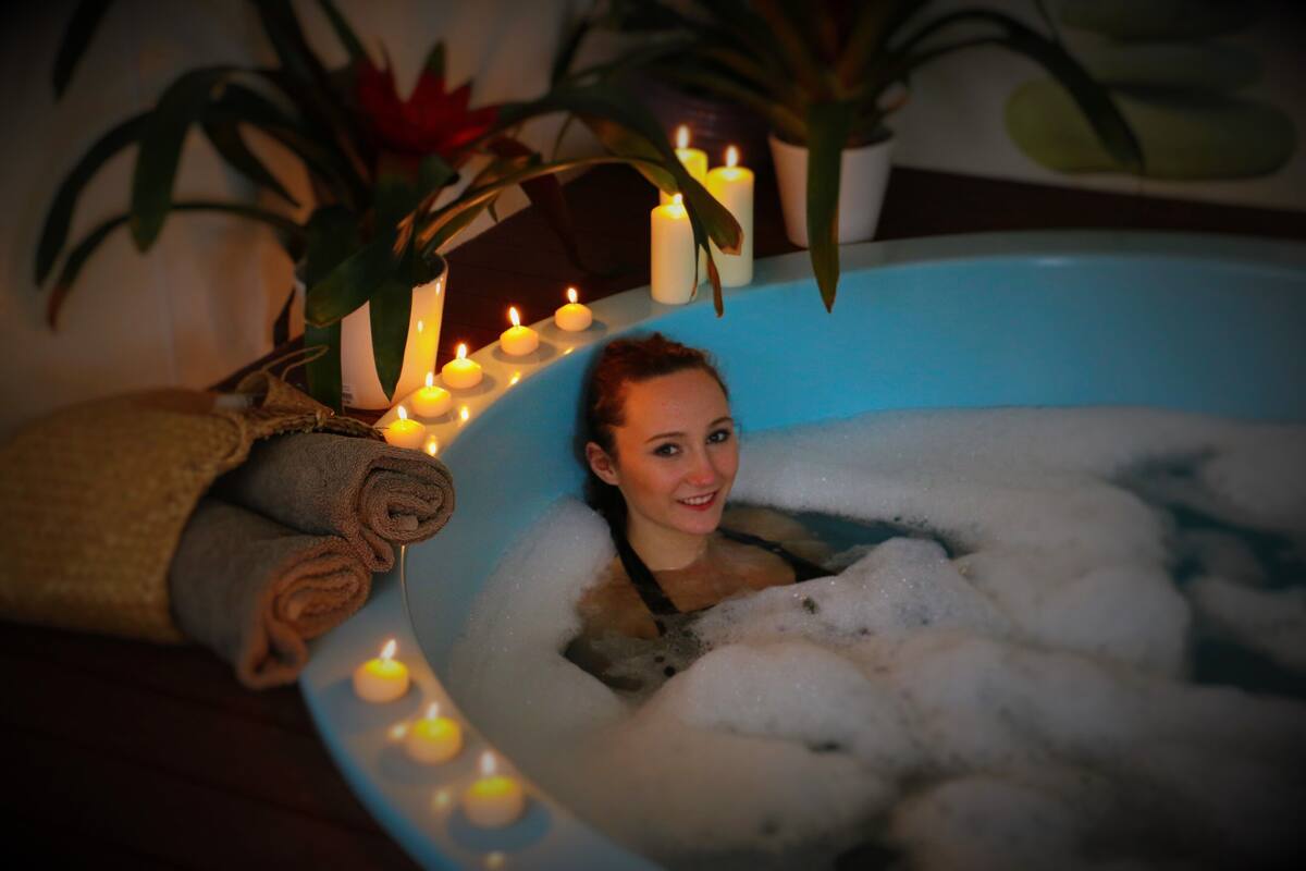 A person in a bathtub filled with bubbles, surrounded by candles and potted plants.