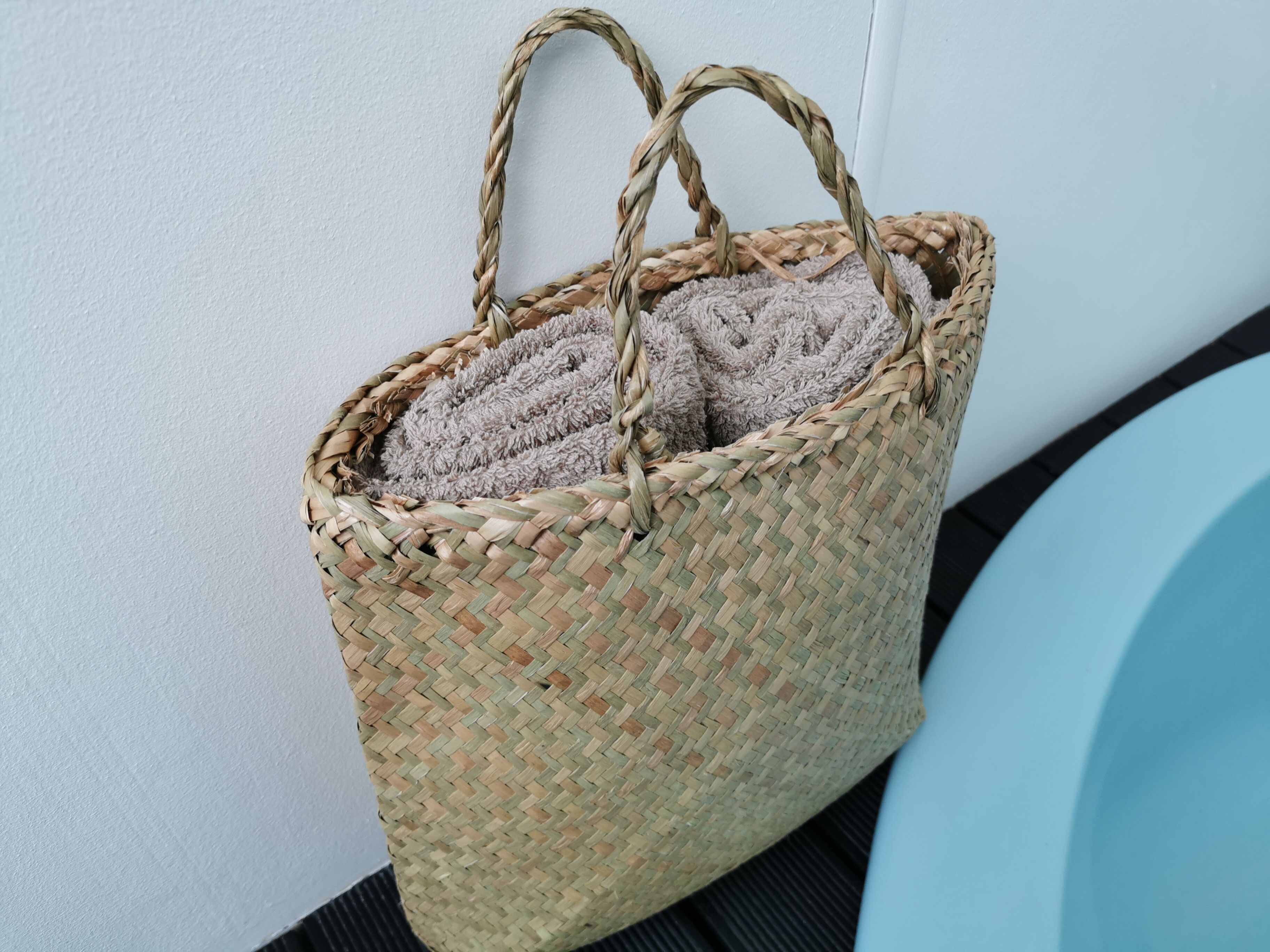 A woven basket with handles, containing folded towels, against a wall.