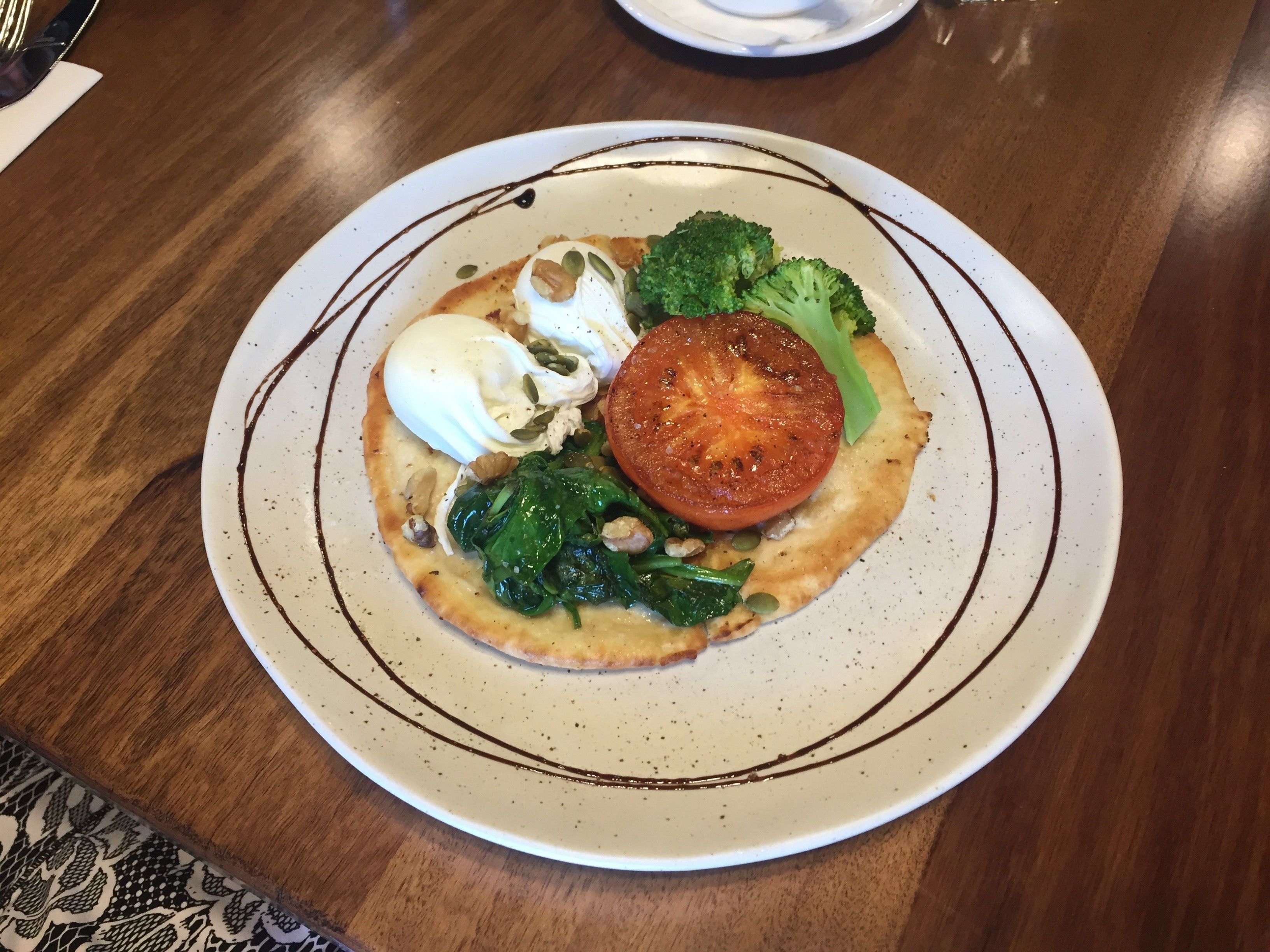 A plate of food with a poached egg, spinach, tomato, and broccoli on a white plate with brown decorative lines.