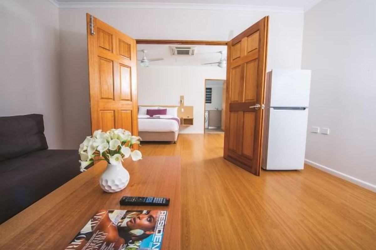 Una stanza con un letto, un frigorifero bianco, un tavolo di legno con un vaso di fiori e una rivista, e un divano scuro.