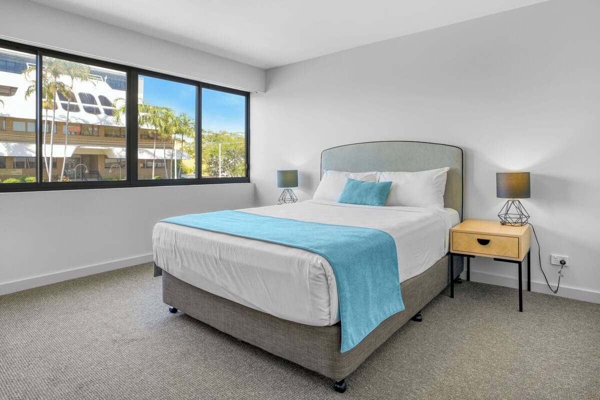 A modern hotel room with a large bed, a nightstand, two lamps, and a view of buildings and trees through large windows.