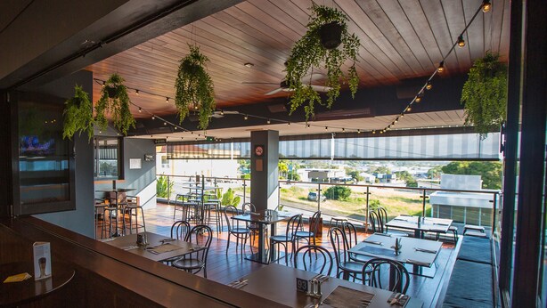 Un comedor interior con mesas y sillas, plantas colgantes y vistas a la ciudad a través de amplios ventanales.