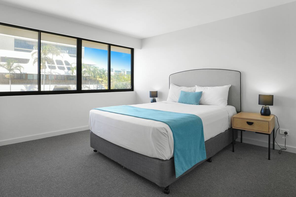 A modern hotel room with a large bed, a nightstand, a lamp, and a view of a building through large windows.