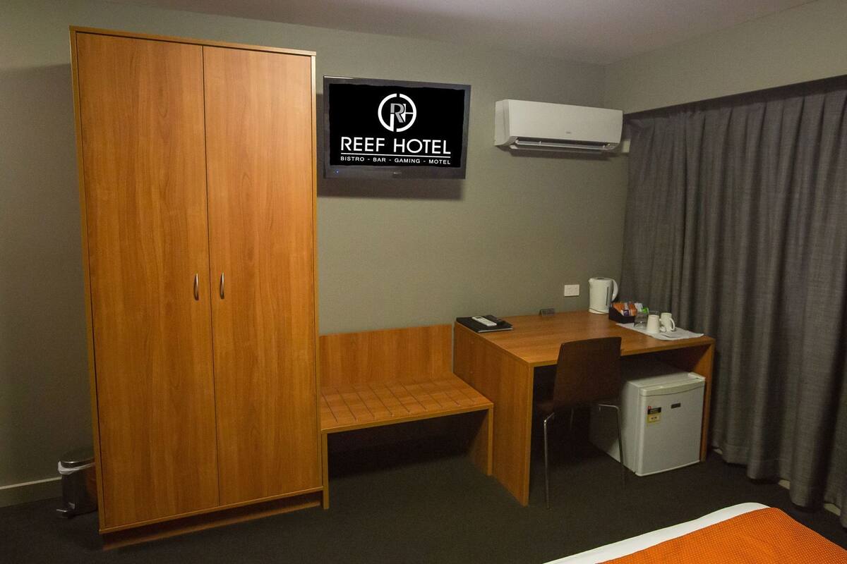 A hotel room with a wardrobe, a desk with a chair, a small fridge, an air conditioner, and a bed with an orange blanket.