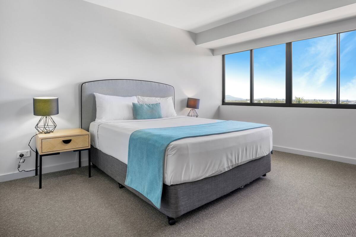 A modern hotel room with a large bed, a nightstand with a lamp, and a view of the city through large windows.