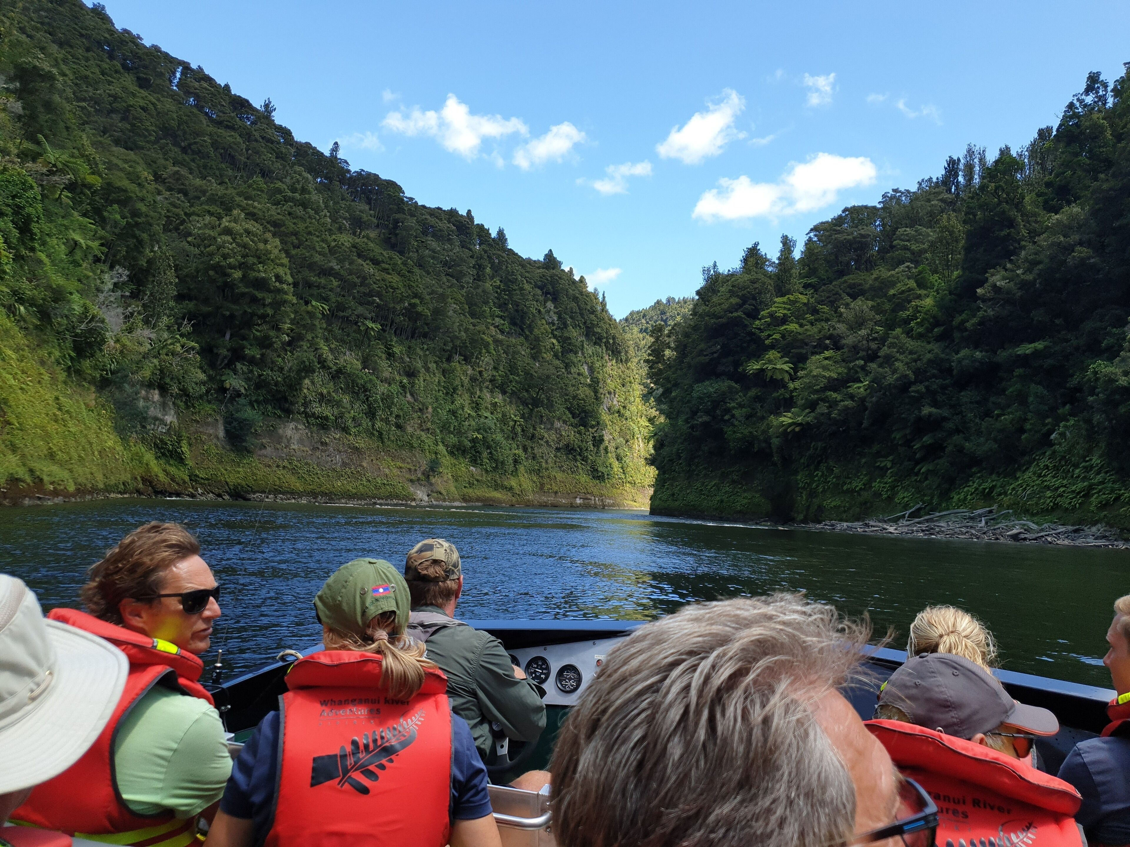 A group of people on a boat, wearing life jackets, surrounded by a river and dense forest.