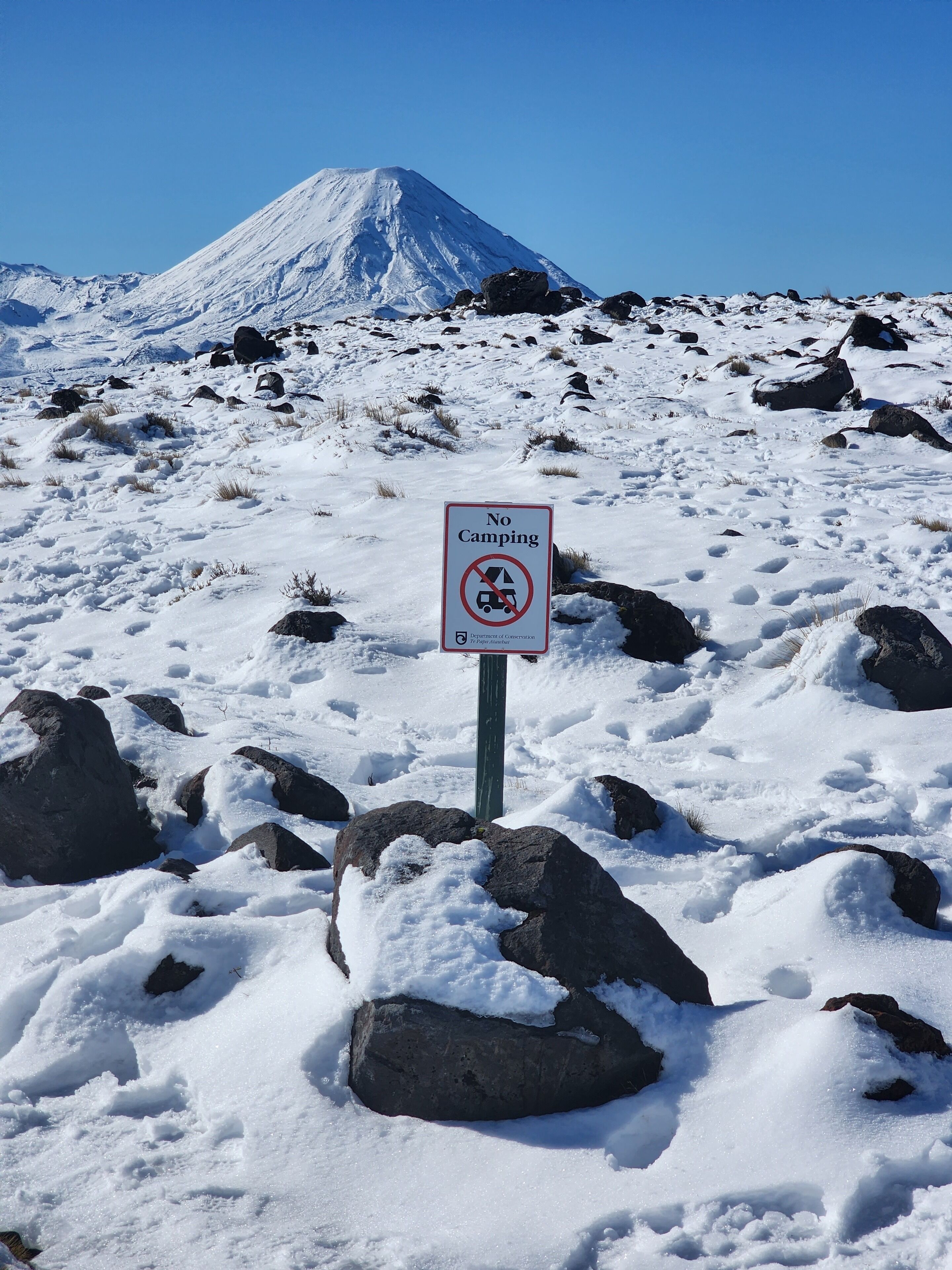 A snowy landscape with a 'No Camping' sign in the foreground and a mountain in the background.
