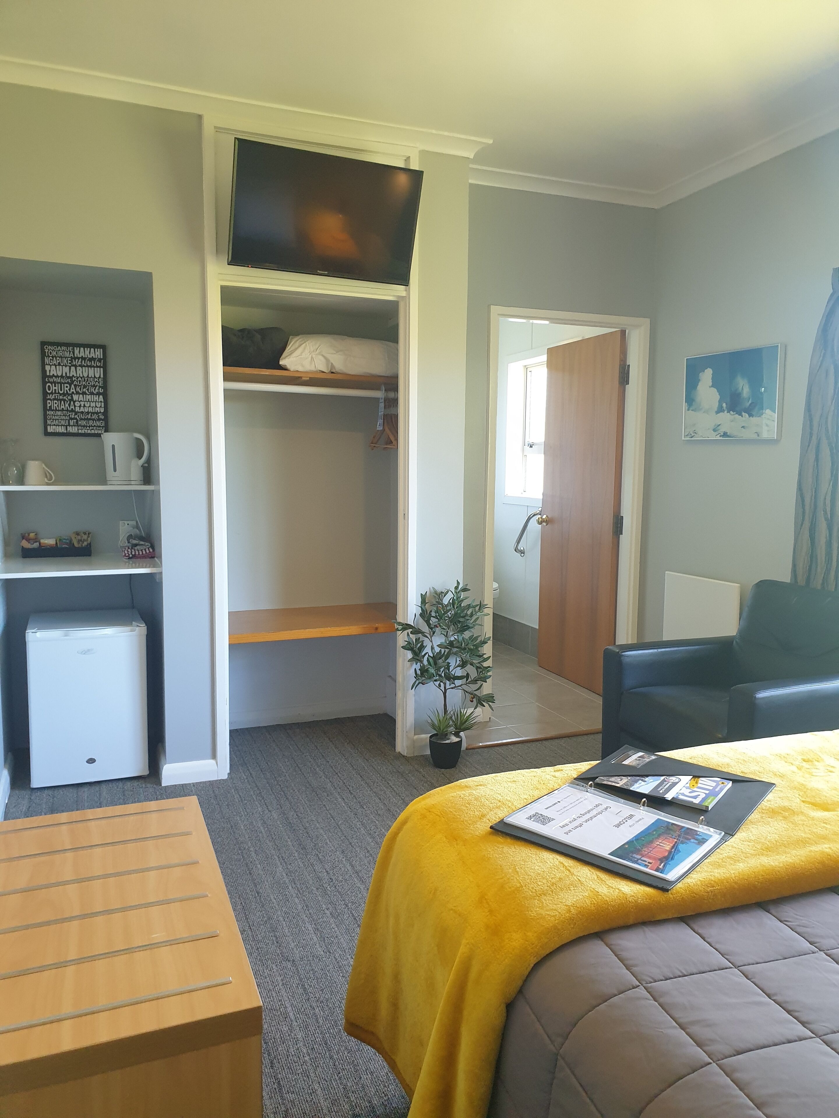 A compact hotel room with a bed, a desk, a small fridge, a potted plant, and a chair.