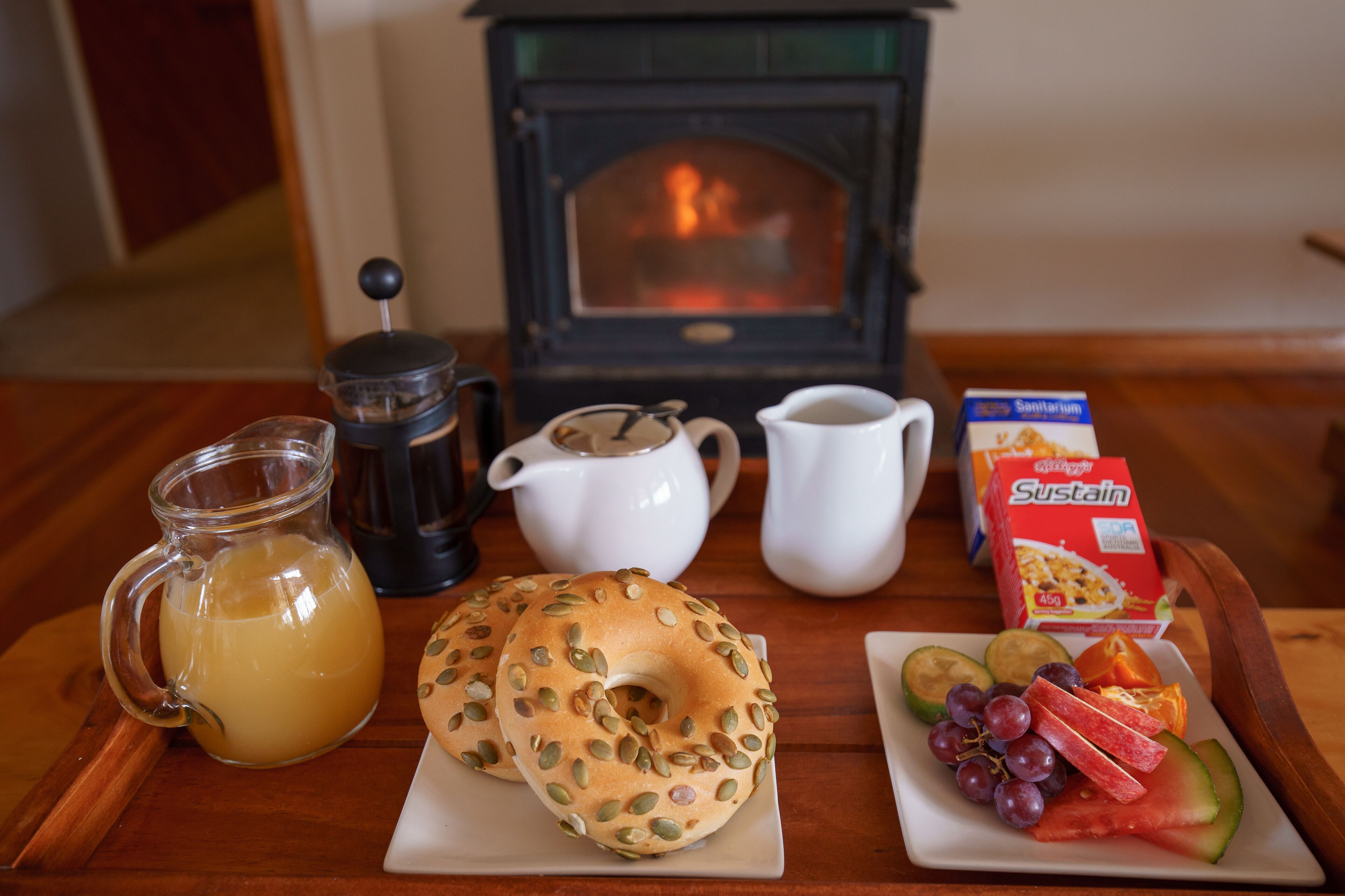 Una preparación para el desayuno que incluye una cafetera, jarra, tetera, bagel y un plato de fruta.