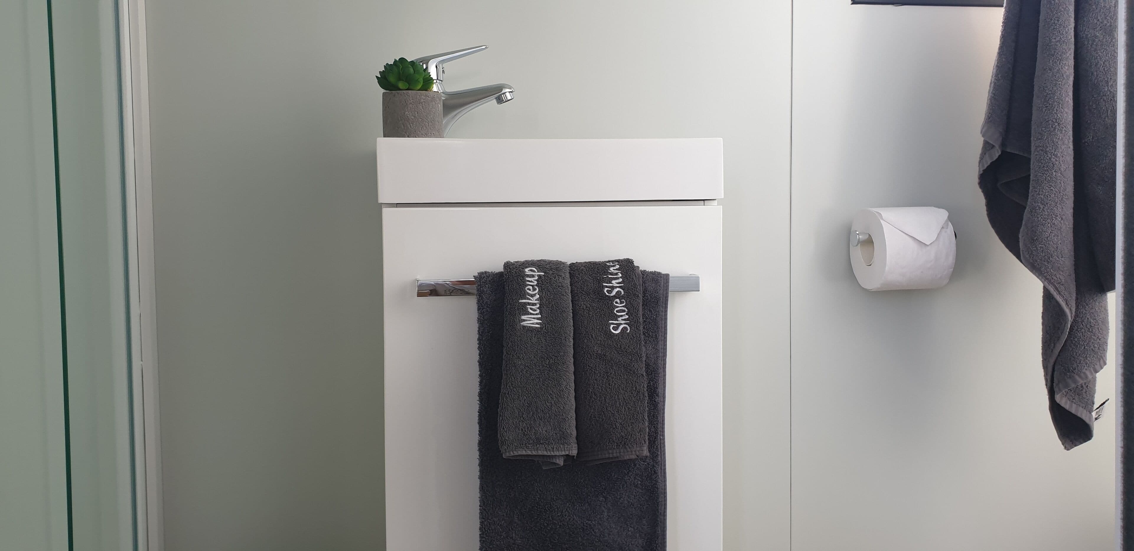 A bathroom with a white sink, a towel rack with two towels, a small potted plant, and a toilet paper holder.