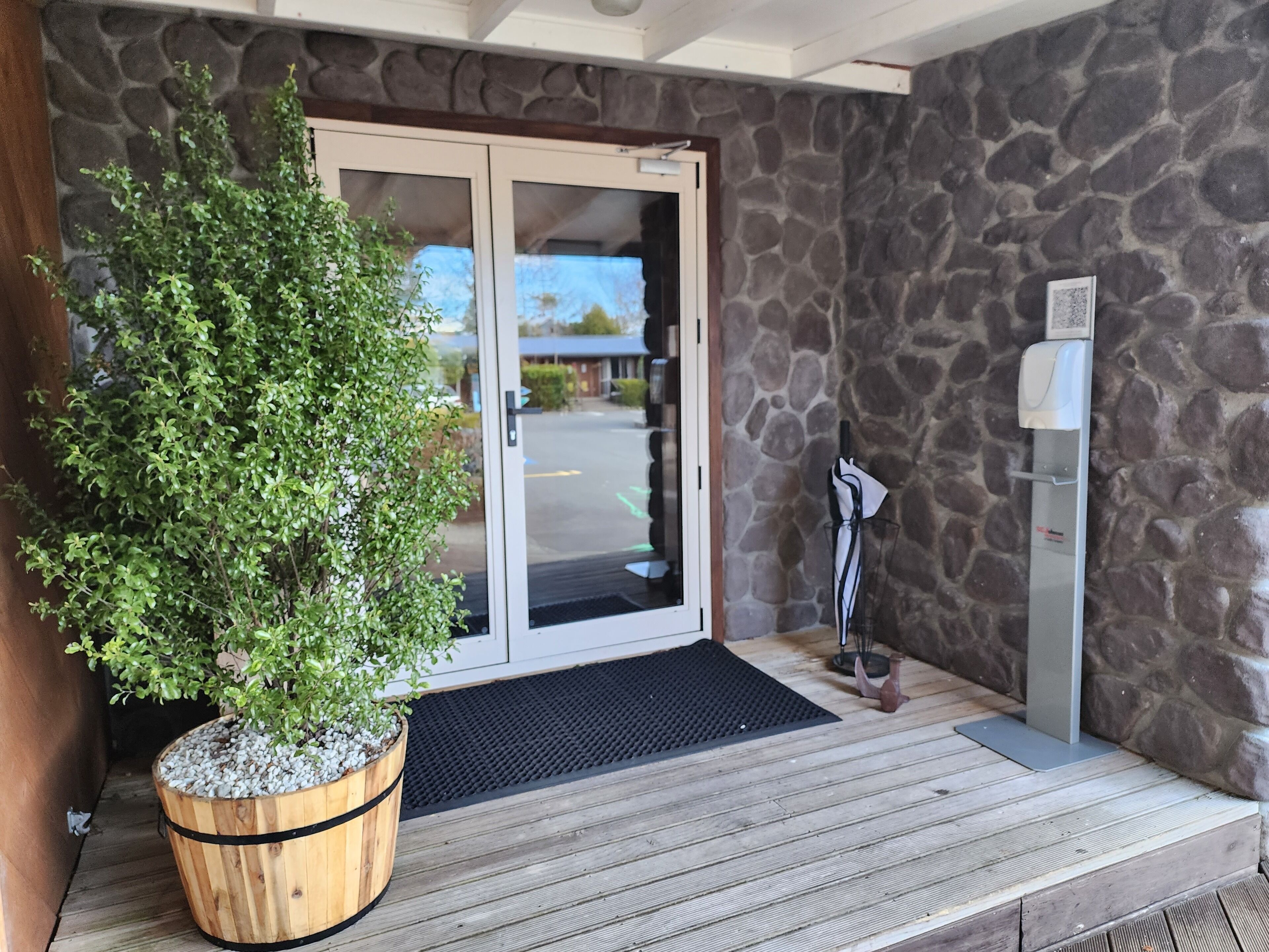 Una jardinera de madera con un arbusto verde, un muro de piedra, una entrada con puerta de cristal, un felpudo negro y un dispensador de agua.