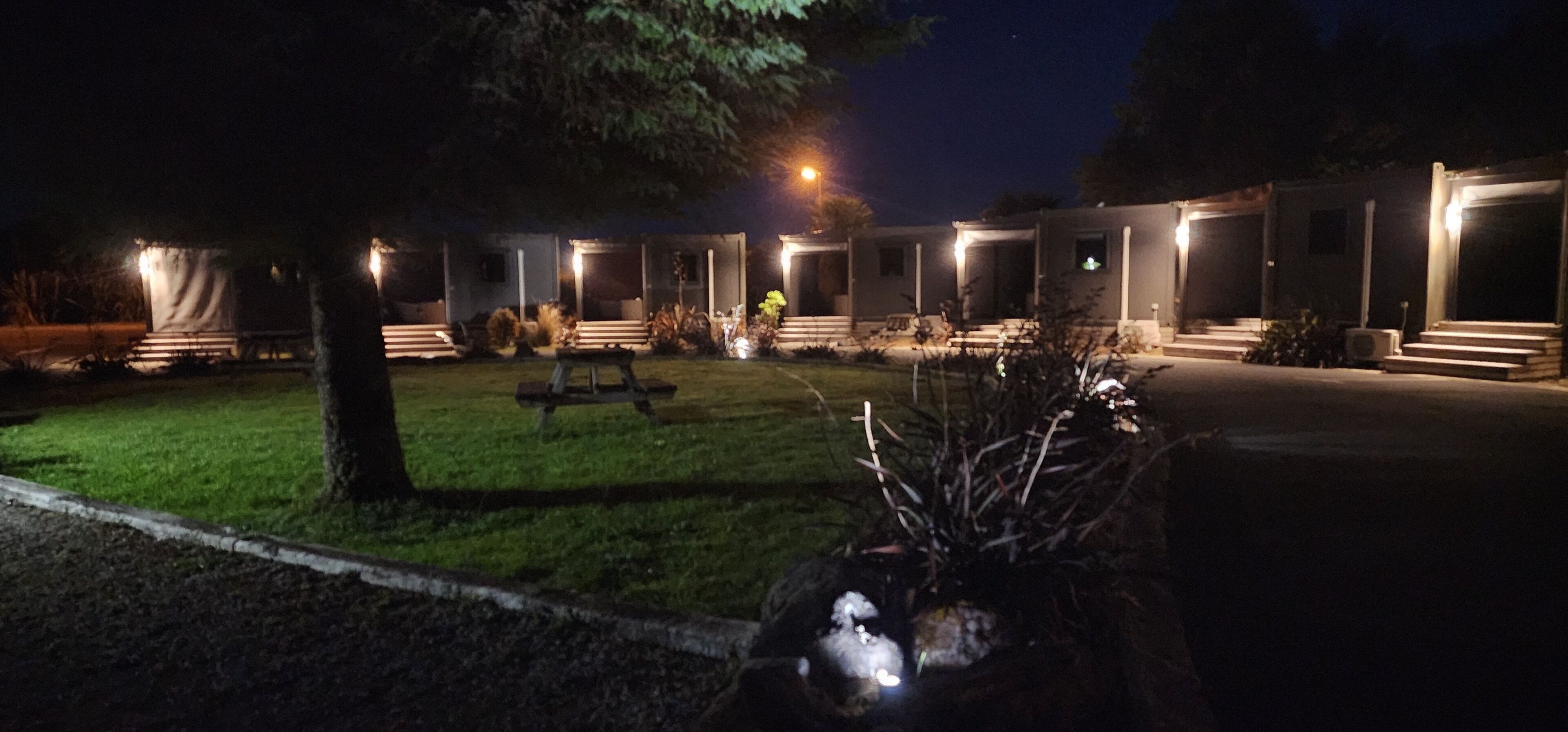 Een nachtzicht van een hotel met meerdere verlichte kamers, een gazon en een bank.