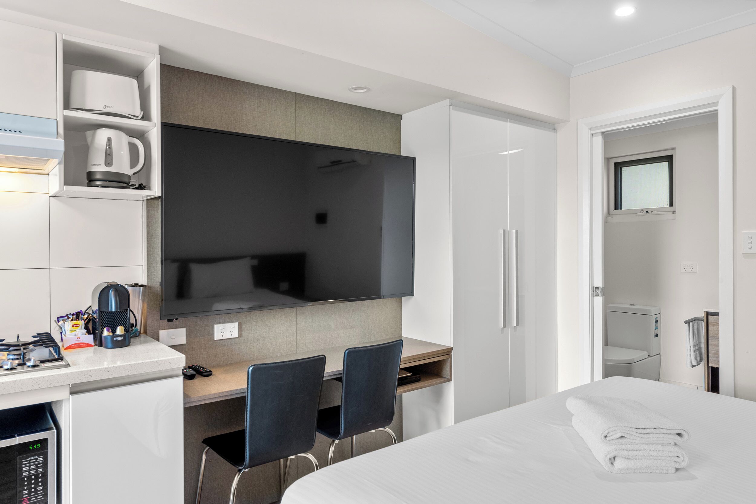 Una habitación de hotel moderna con una televisión de pantalla plana, un escritorio con cafetera, dos sillas, una cama con una toalla blanca doblada con cuidado y un vestidor.