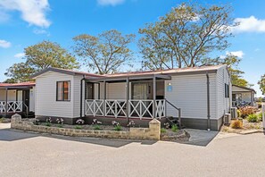 A single-story mobile home with a porch, a small garden with flowers, and a clear blue sky in the background.