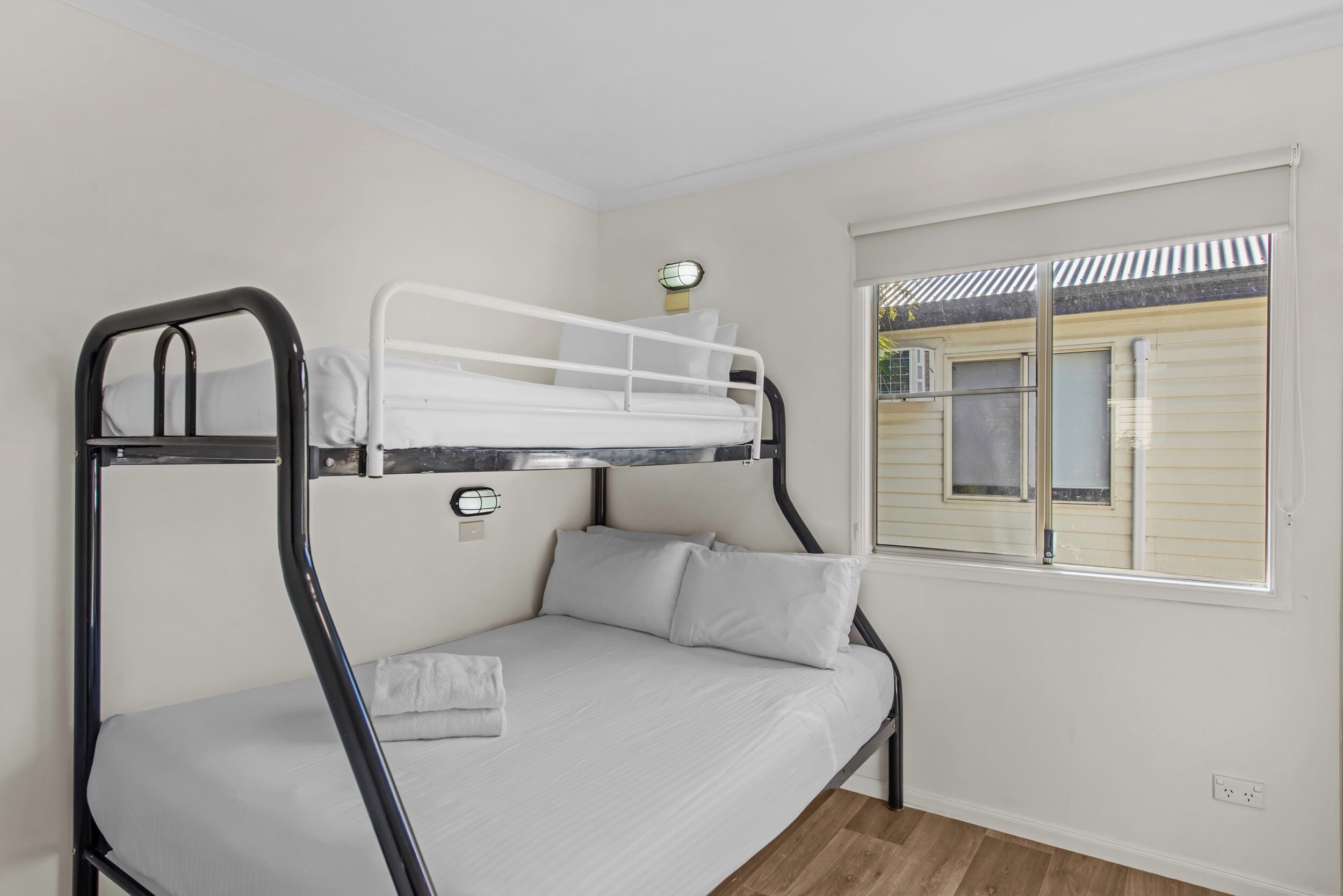 Un lit superposé avec un matelas et des oreillers blancs, une petite table avec une lampe et une fenêtre avec des stores.