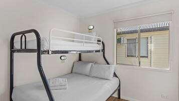Un lit superposé avec un matelas et des oreillers blancs, une petite table avec une lampe et une fenêtre avec des stores.
