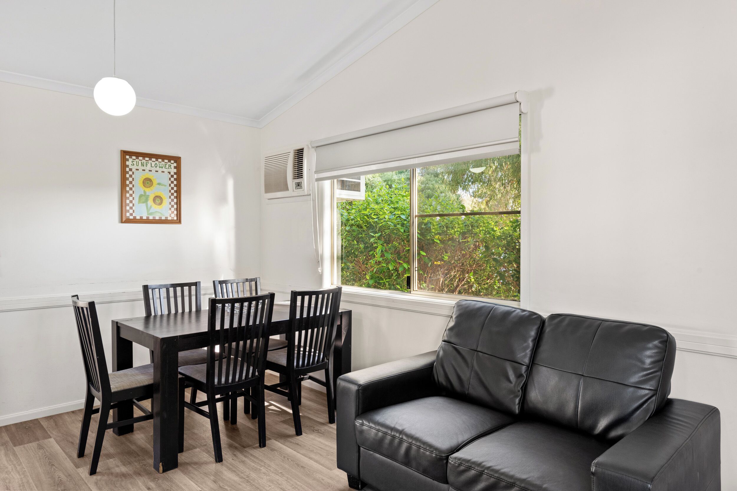 A dining area with a table and chairs, a black leather sofa, a window with a view of greenery, and a framed picture on the wall.