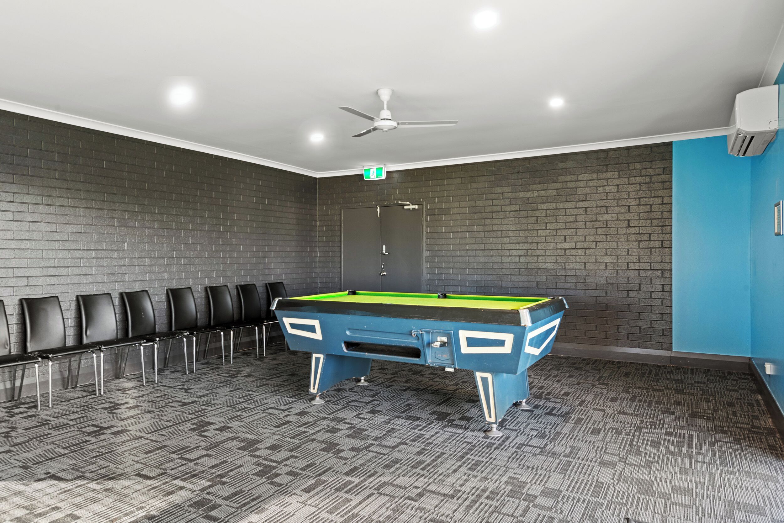 A billiards room with a pool table, chairs, a ceiling fan, and a door marked with an exit sign.