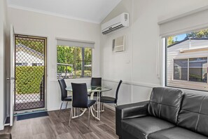 A modern room with a glass table, four chairs, a black leather sofa, and a view of greenery outside.