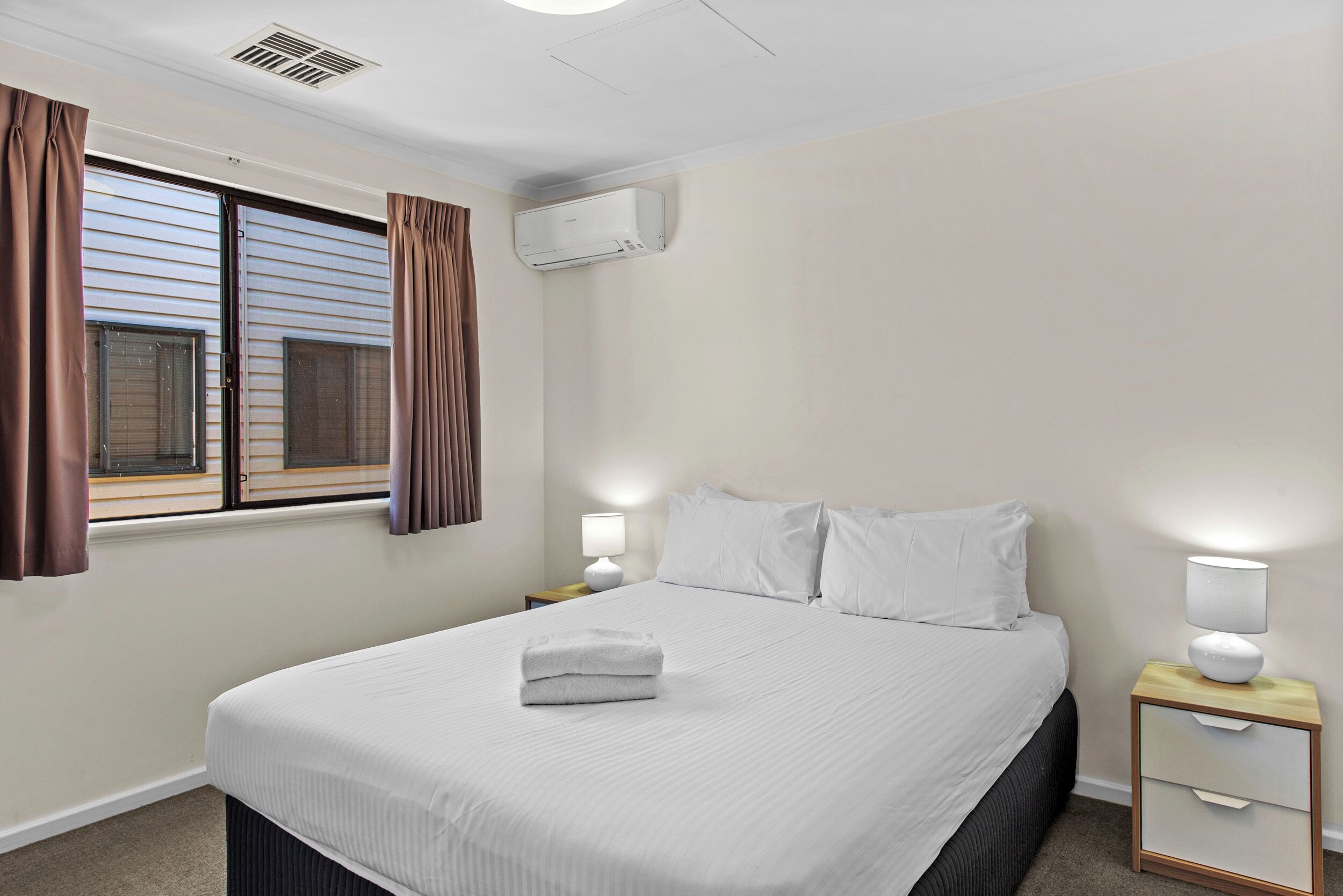 Uma cama bem arrumada com lençóis brancos, um abajur e uma janela com cortinas.