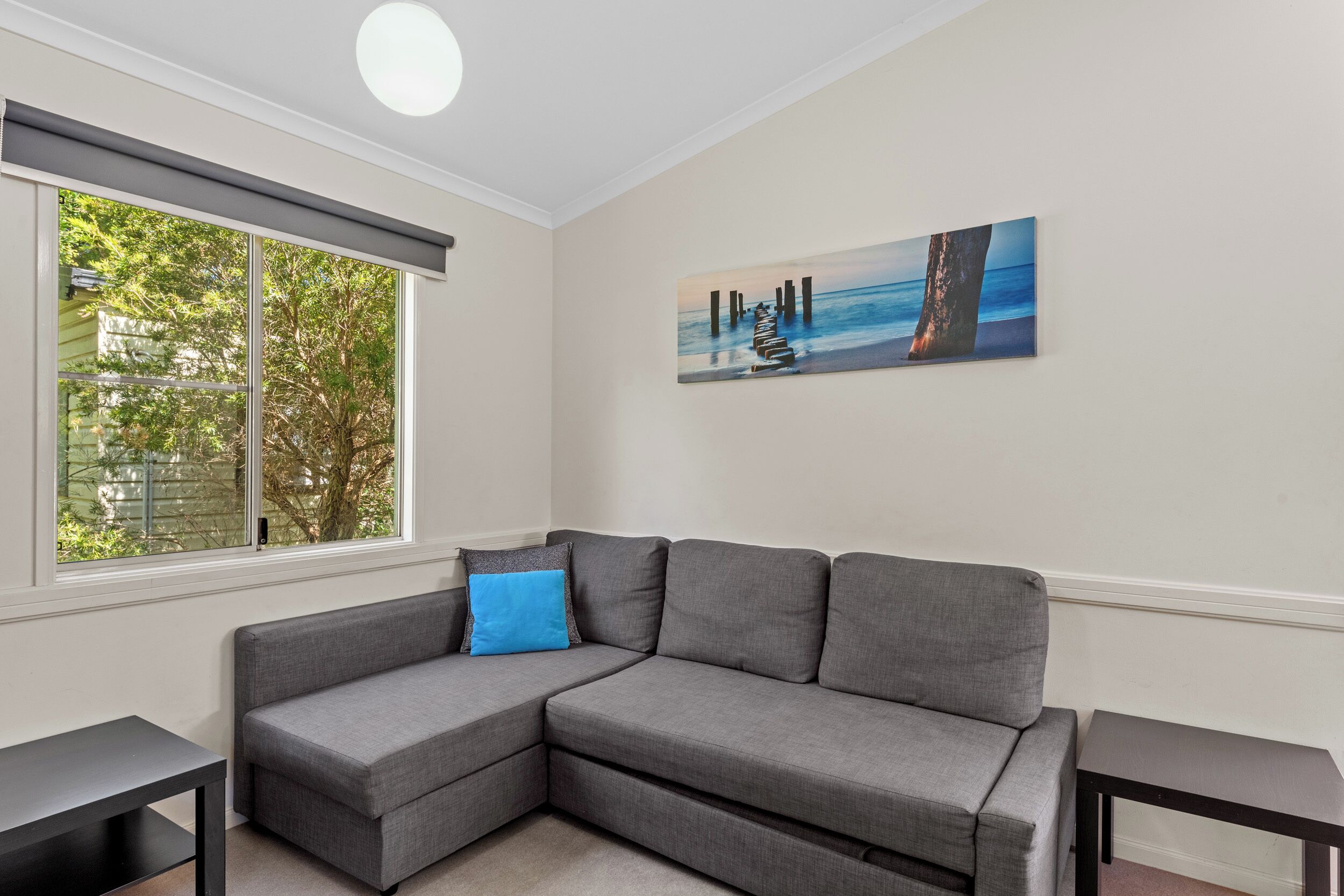 A modern living room with a gray sectional sofa, a blue accent pillow, a painting of a pier, and a large window with a view of trees.
