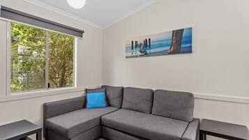 A modern living room with a gray sectional sofa, a blue accent pillow, a painting of a pier, and a large window with a view of trees.