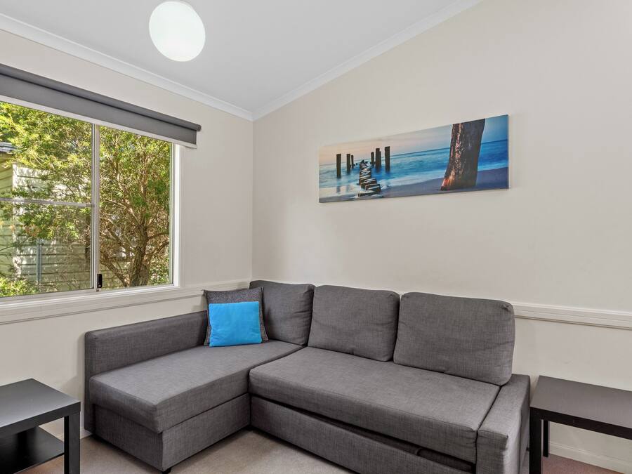 A modern living room with a gray sectional sofa, a blue accent pillow, a painting of a pier, and a large window with a view of trees.