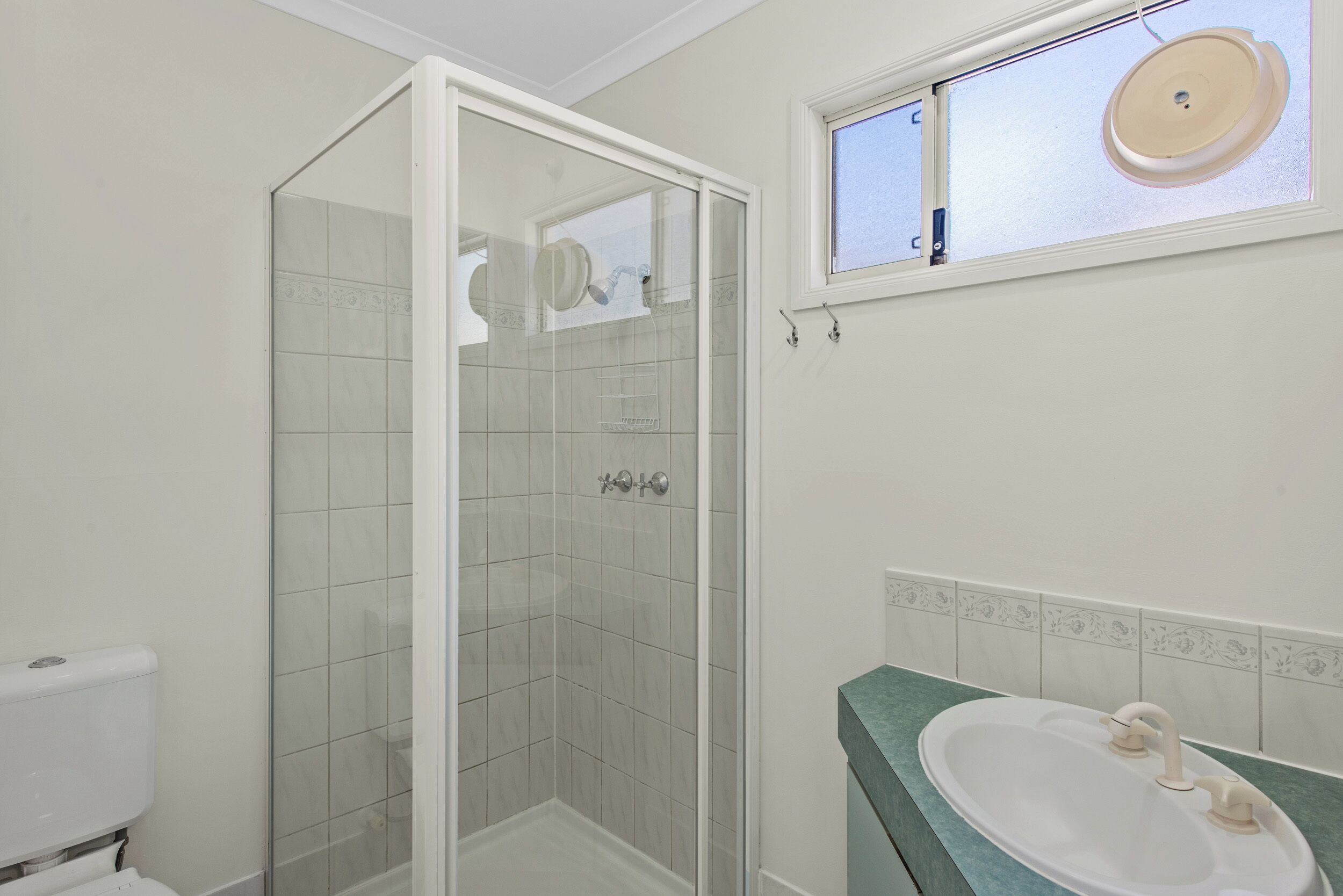 Un baño con ducha envuelta en vidrio, un lavamanos con encimera verde y una ventana sobre el lavamanos.