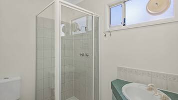 Un baño con ducha envuelta en vidrio, un lavamanos con encimera verde y una ventana sobre el lavamanos.