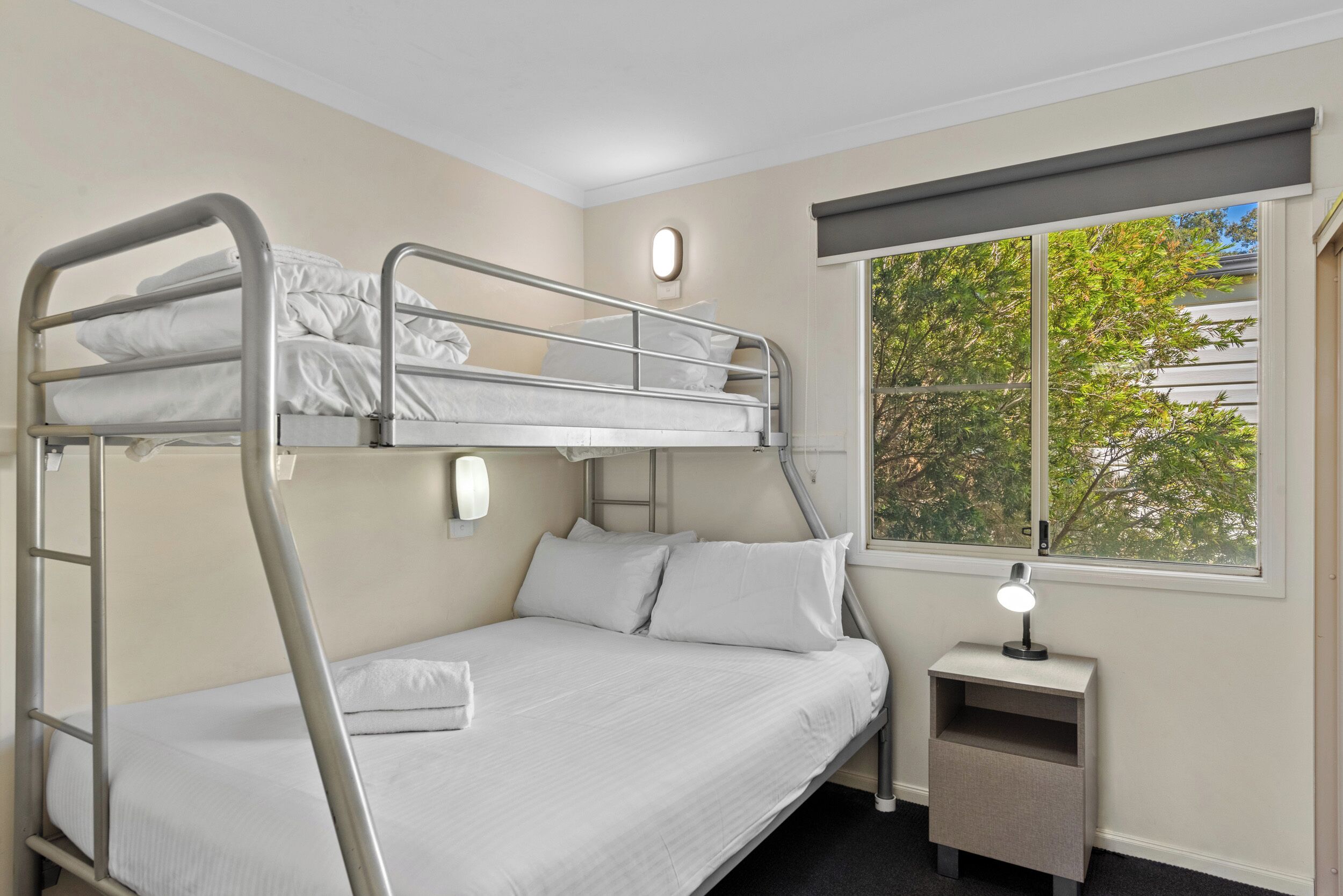 A bunk bed room with a small bedside table and a window with a view of trees.