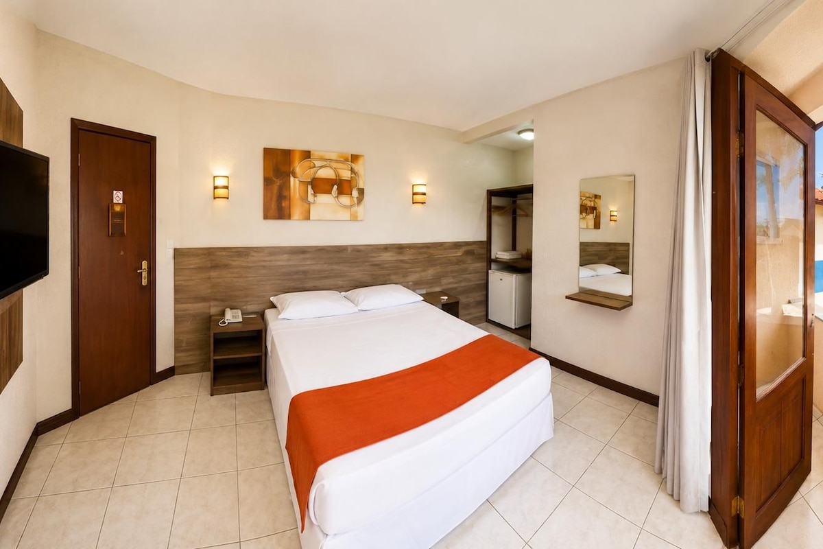 Quarto de hotel com cama, escrivaninha, telefone, televisão e espelho.