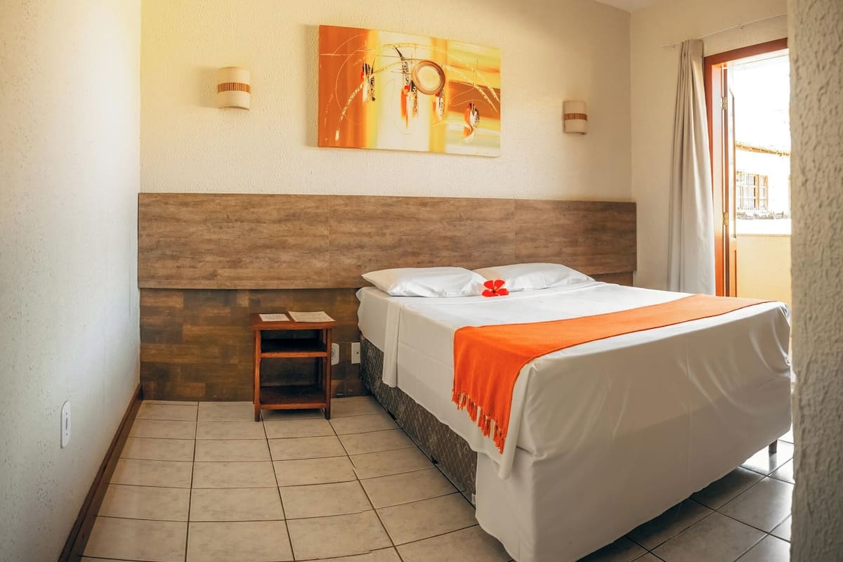 Une chambre d’hôtel avec un lit, une petite table de chevet en bois, un tableau accroché au mur et une fenêtre avec des rideaux.