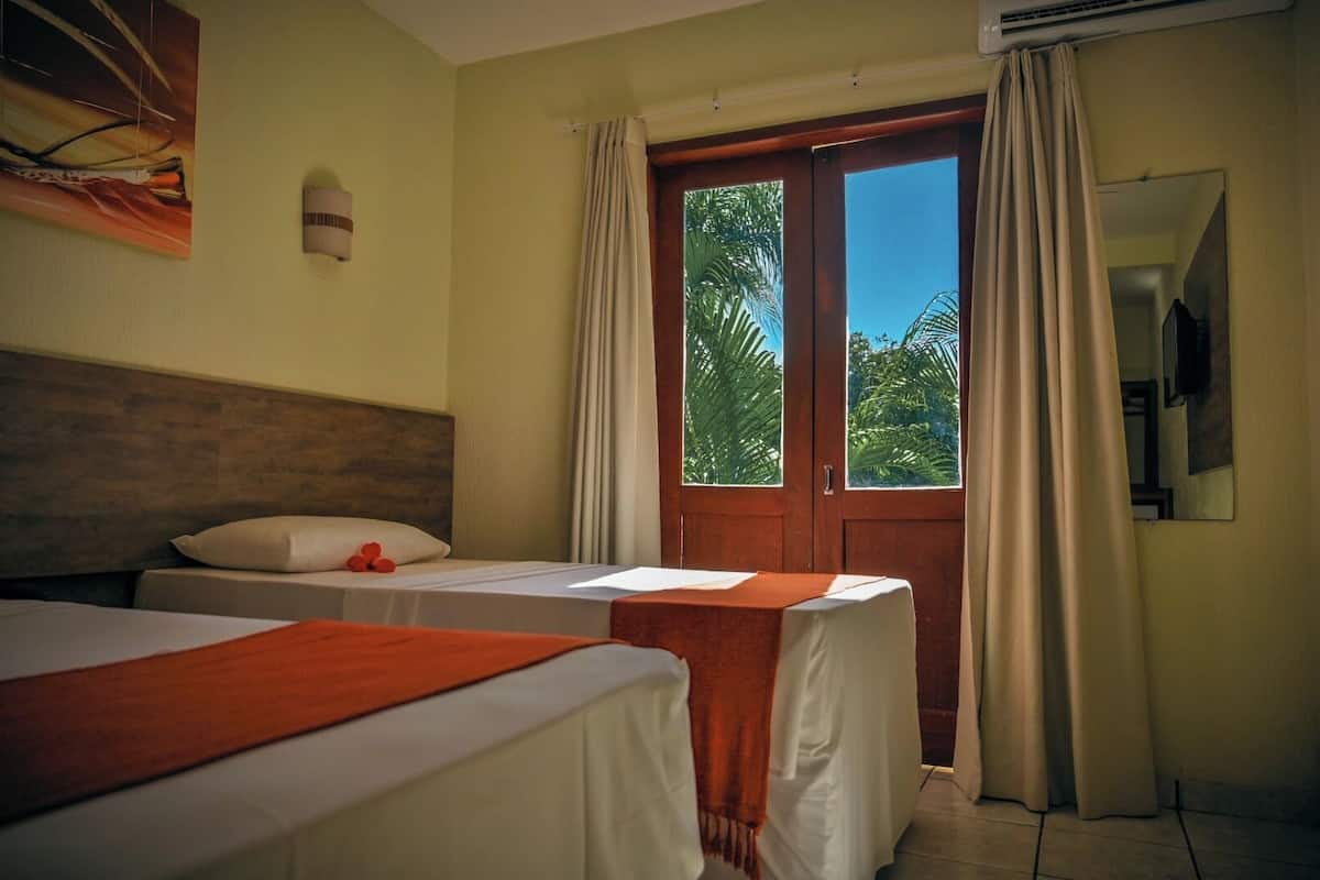 Une chambre d’hôtel avec deux lits, une tête de lit en bois, une grande fenêtre avec des rideaux et un appareil de climatisation.