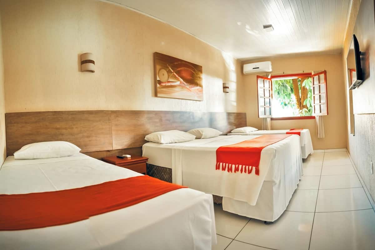 Une chambre d’hôtel avec trois lits, un tableau au mur, une fenêtre donnant sur un paysage verdoyant et un appareil de climatisation.