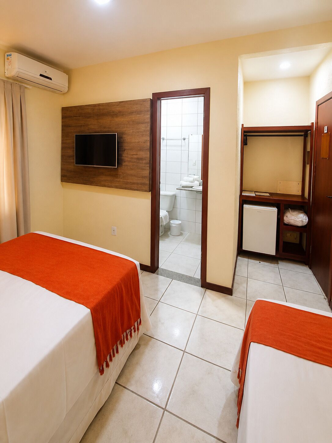 A hotel room with two beds, a TV mounted on the wall, a small desk with a mirror, and a bathroom visible through an open door.