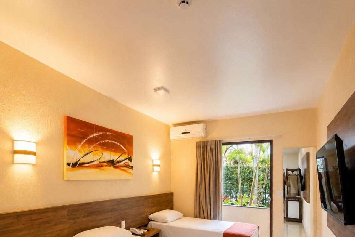 A hotel room with two beds, a painting on the wall, a TV, and a view of greenery outside.
