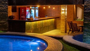 A bar area with a Coca-Cola refrigerator, a bottle of wine, and a coffee machine. There is a pool with lights around it and a seating area with chairs. - Quinta do Sol Praia Hotel (Porto Seguro)