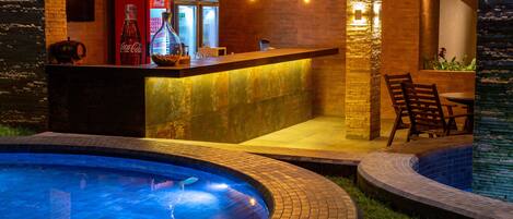 Una zona de bar con un refrigerador Coca-Cola, una botella de vino y una cafetera. Hay una piscina con luces y un área de descanso con sillas.