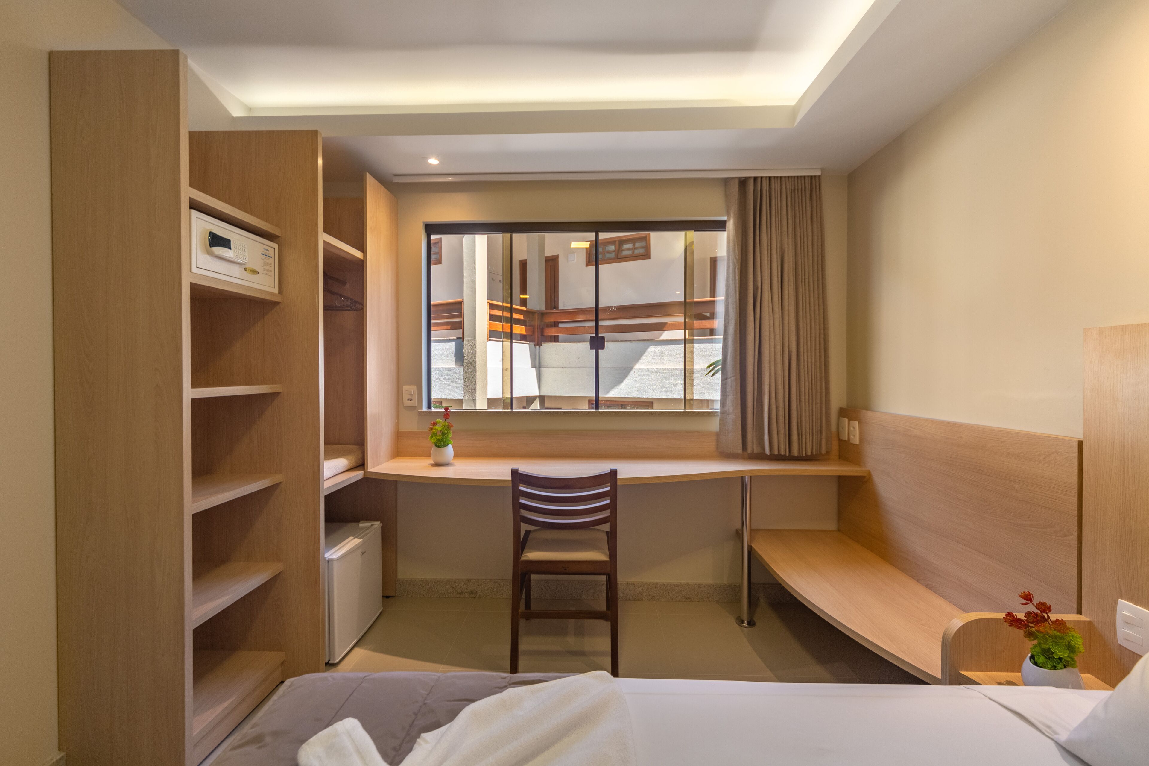 A compact hotel room with a bed, a desk with a chair, a small radiator, a window with curtains, and a wooden wardrobe.