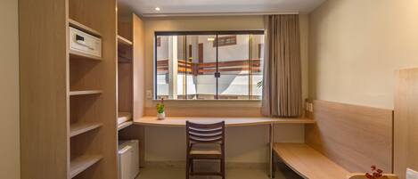 A compact hotel room with a bed, a desk with a chair, a small radiator, a window with curtains, and a wooden wardrobe.
